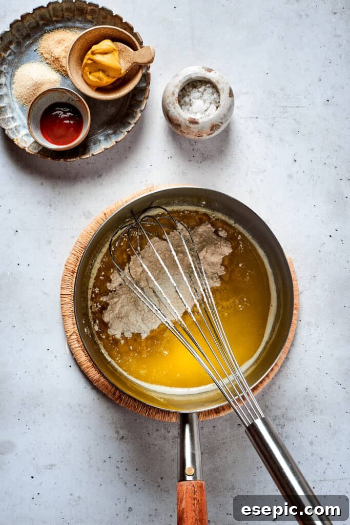 Master the art of homemade soft pretzels and pair them with our decadent, mustard-infused cheese dip. A metal whisk expertly blends a creamy, golden cheese sauce in a stainless steel pot, ensuring a smooth, lump-free consistency.