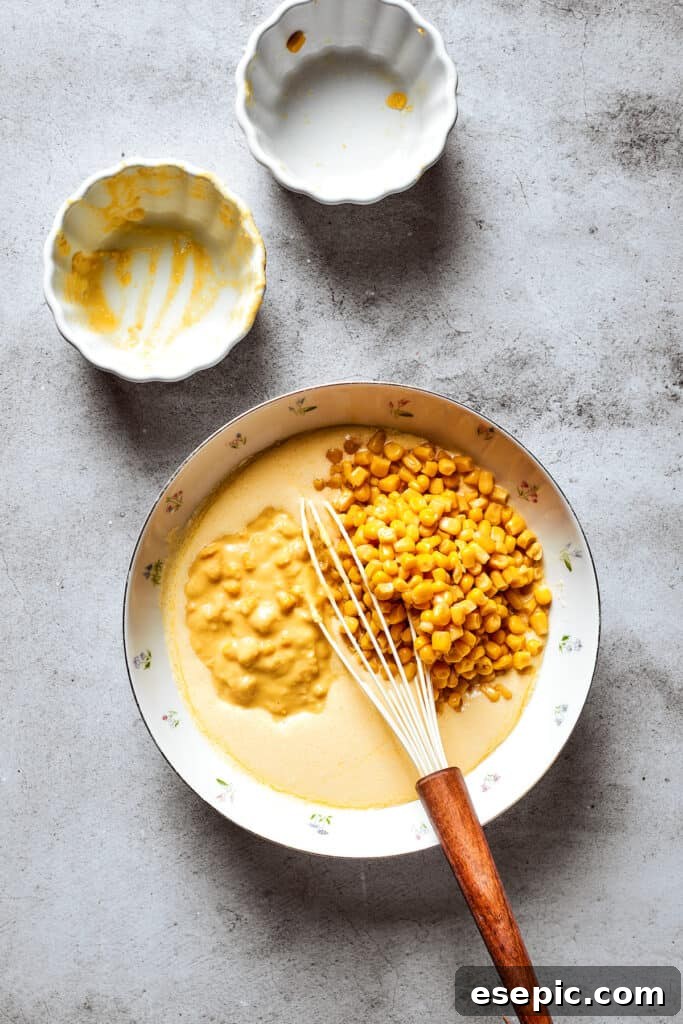 Whole kernel corn and creamed corn are being added to a large mixing bowl containing the whisked souffle base.