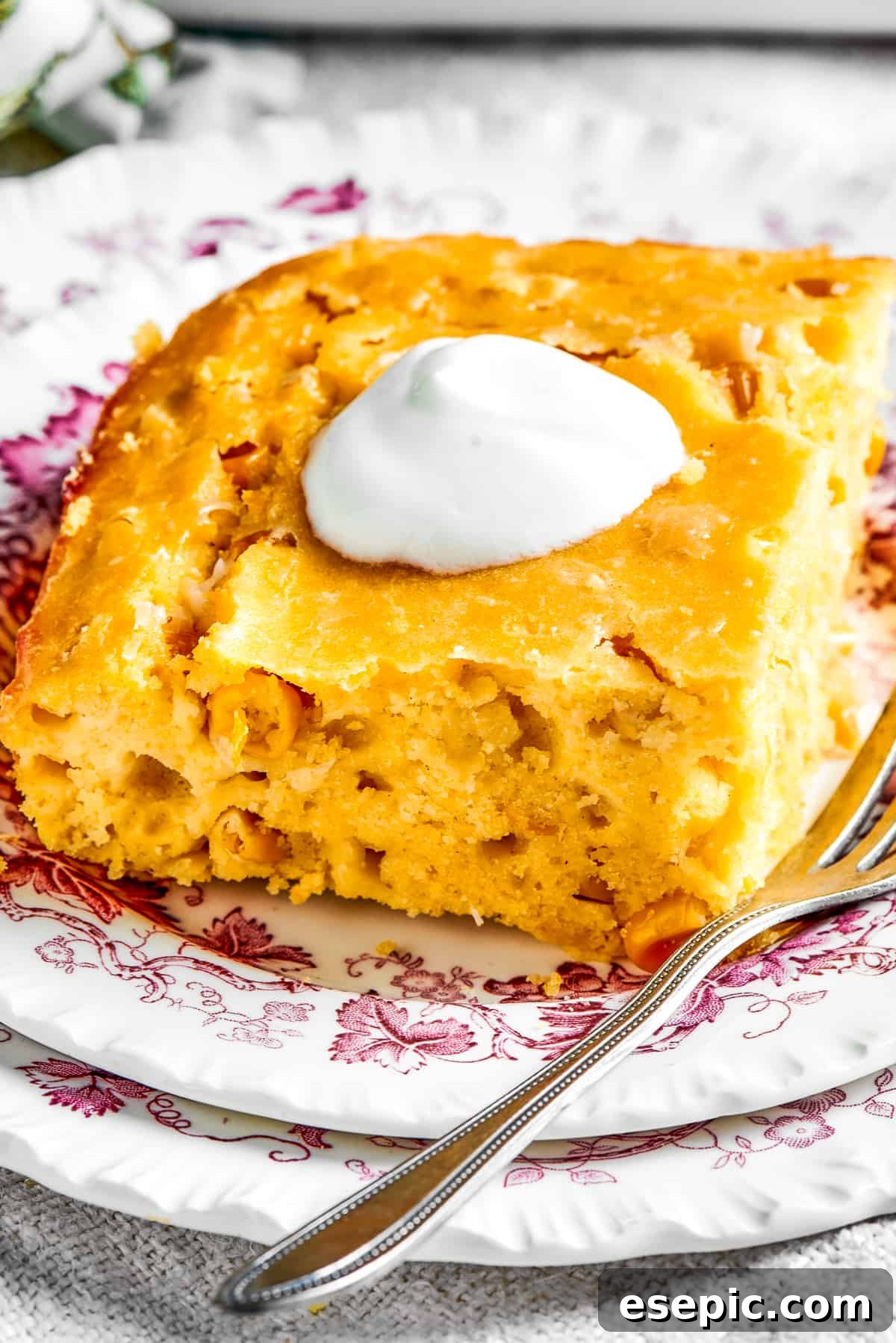 A square of golden corn souffle elegantly served on a white dish and garnished with a dollop of sour cream and a sprig of fresh herb.