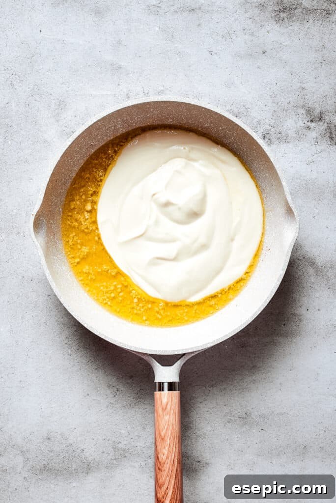 Cream cheese cubes and heavy cream are added to the melted butter and garlic in a white skillet, ready to be whisked into a sauce.