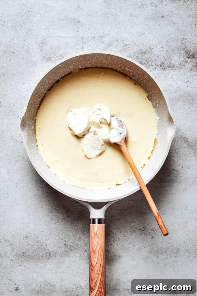 A wooden spoon stirs softened cream cheese into a creamy sauce in a white skillet until smooth.