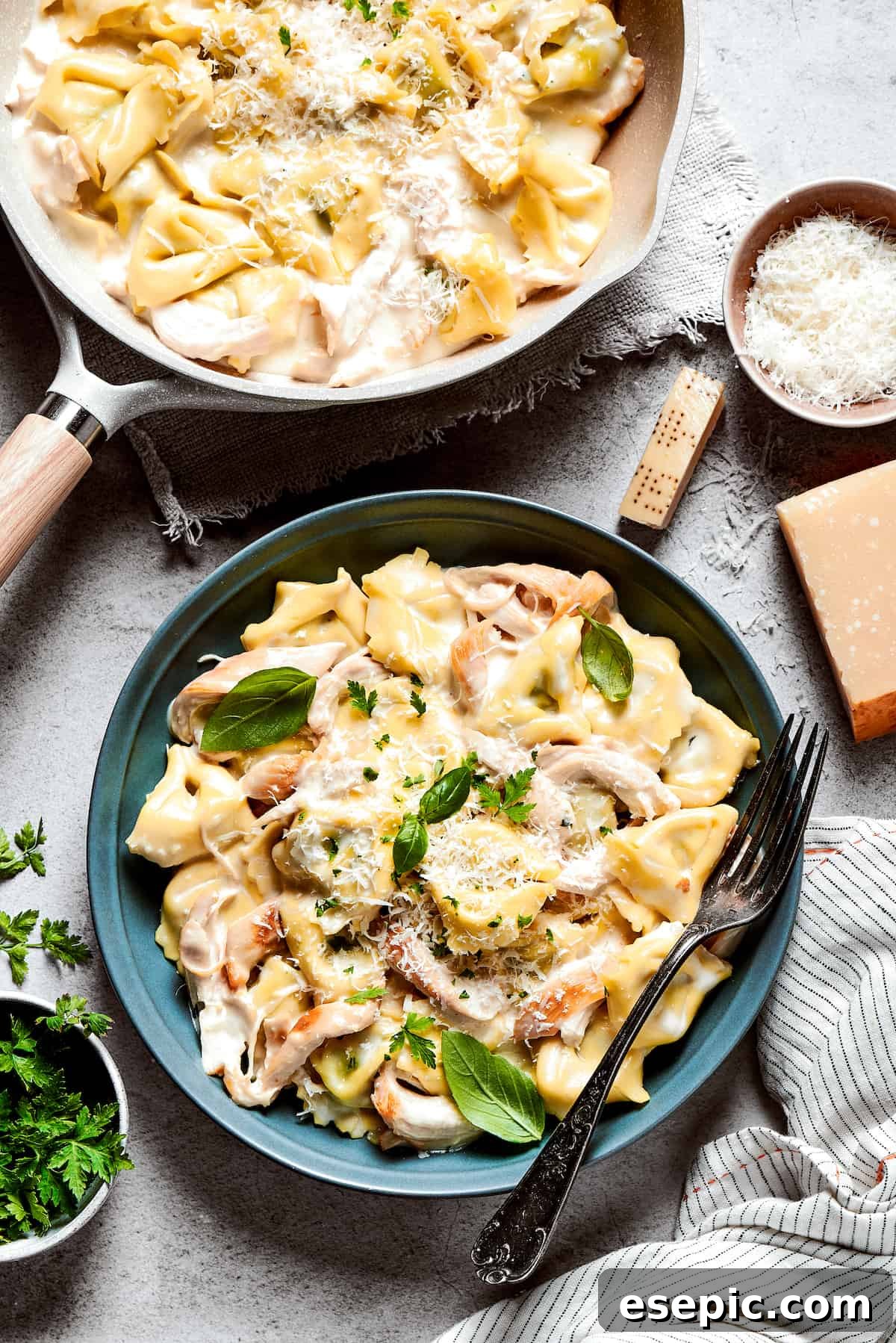 A beautiful blue bowl of creamy chicken tortellini alfredo with a fork resting on the edge, a skillet of the pasta dish sits in the background.