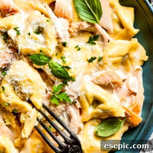 A blue bowl with a fork holds a creamy portion of chicken tortellini garnished with parmesan cheese and basil leaves.