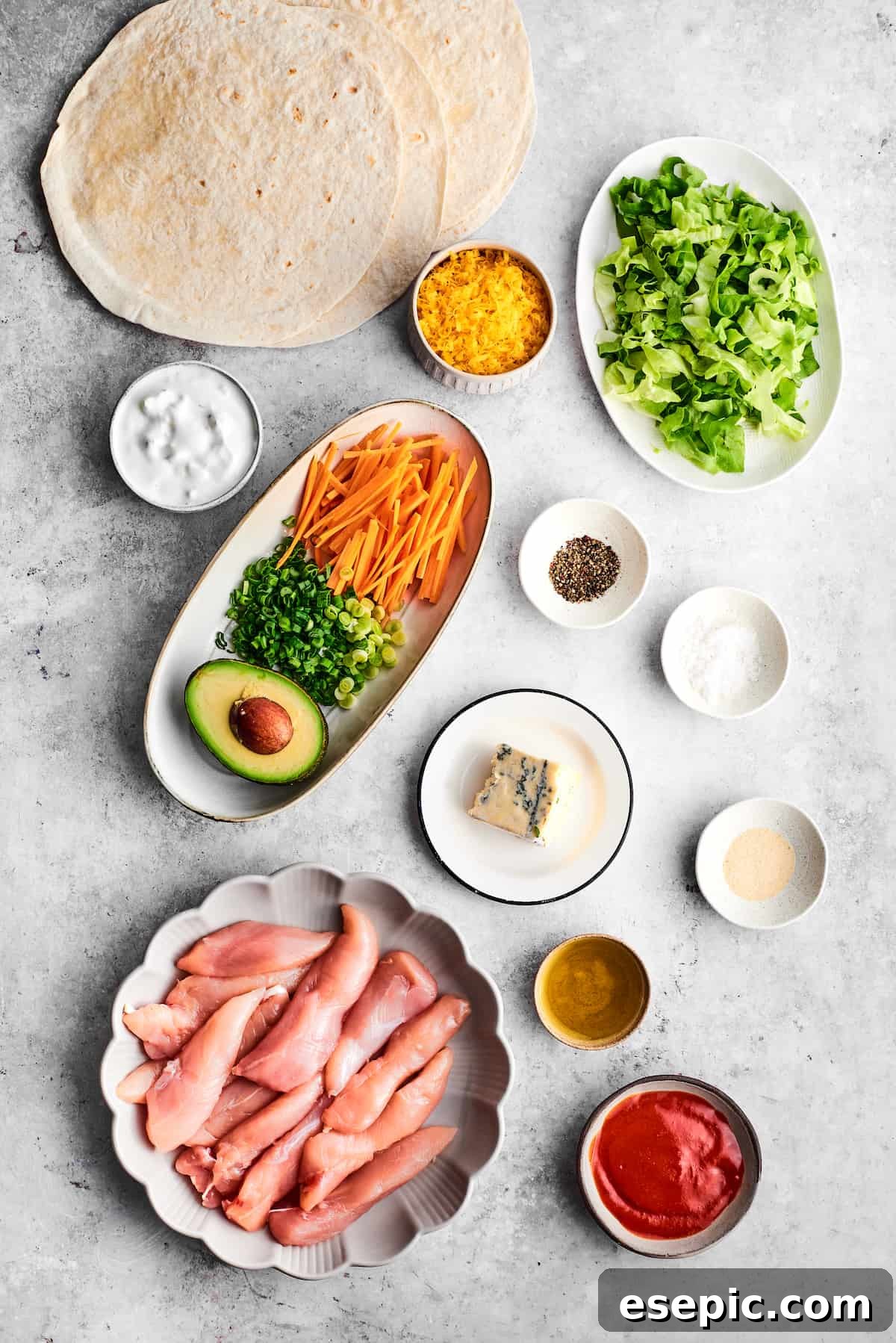 The array of fresh ingredients laid out for making Buffalo Chicken Wraps: seasoned chicken tenders, olive oil, salt and pepper, garlic powder, hot sauce, ranch dressing, avocado, green onions, carrots, tortillas, shredded cheddar cheese, and crumbled blue cheese.