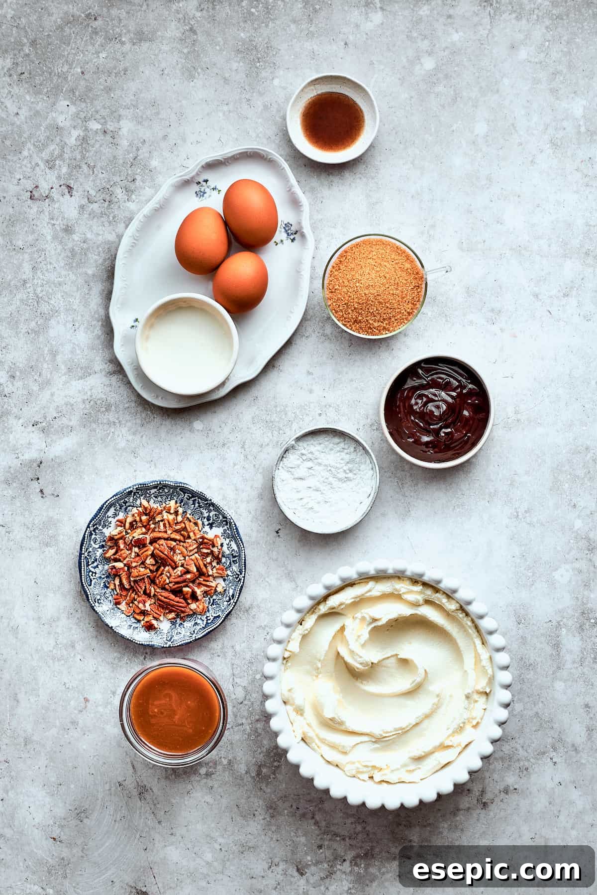 The ingredients for turtle cheesecake are shown portioned out on a white background: eggs. pecans, caramel, cream cheese, chocolate chips, brown sugar, vanilla, sour cream.