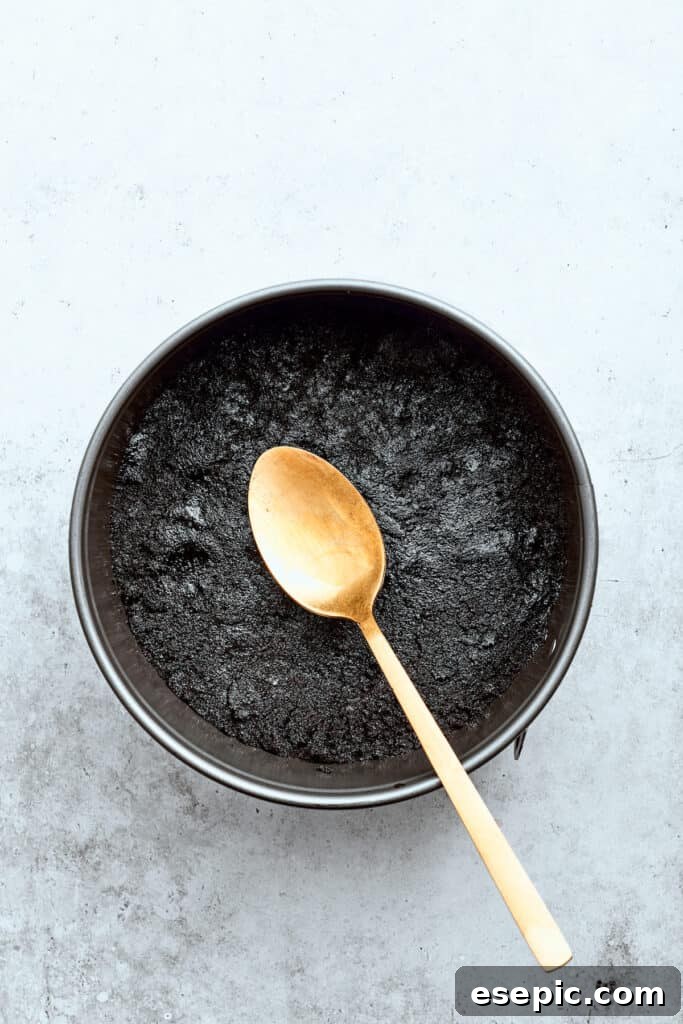 A spoon pats a chocolate cookie crust into a springform baking pan.
