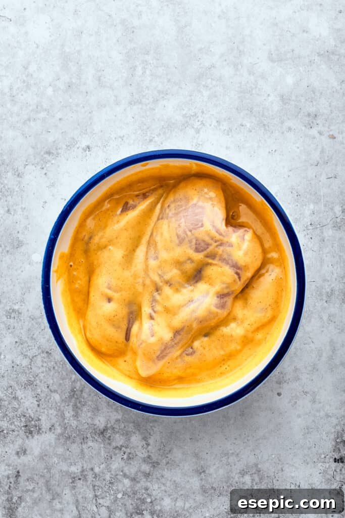 Chicken breasts marinate in a white bowl of yellow honey mustard sauce.