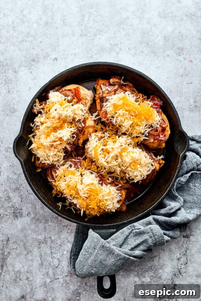 Four chicken breasts are topped with bacon and cheese in a cast iron skillet.