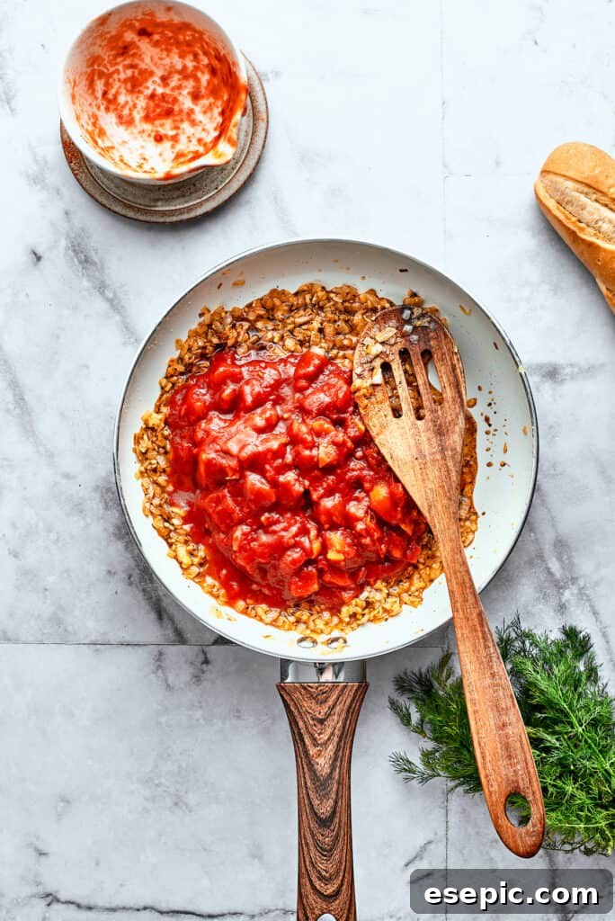 Thick diced tomatoes are stirred into a white skillet with a wooden spoon.