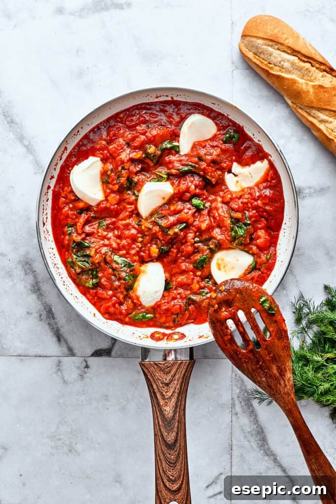 Slices of fresh mozzarella cheese are seen peeking through a hearty red sauce in a skillet with a wooden spoon.