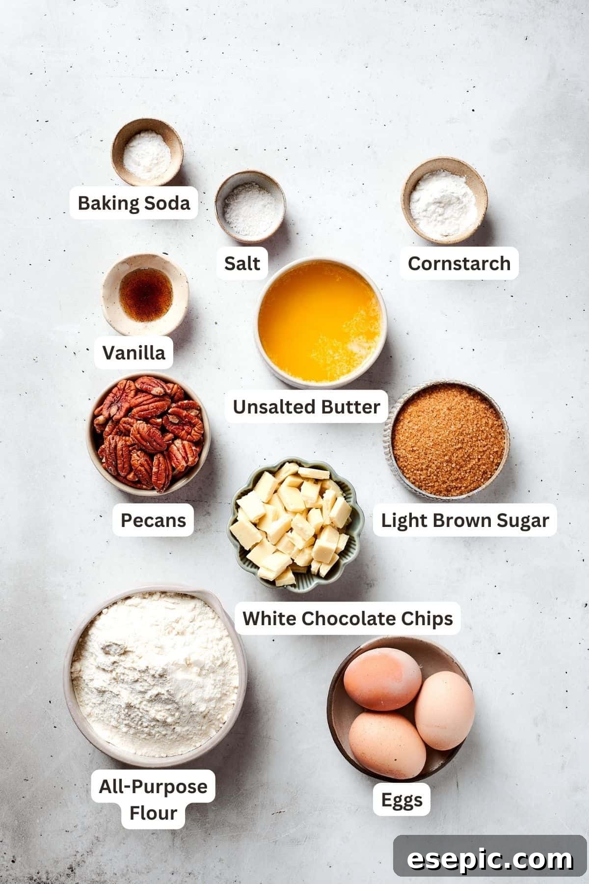 The organized ingredients for making blonde brownies are neatly arranged on a counter. They include a bowl of flour, cornstarch, baking soda, and salt, along with separate containers for vanilla extract, brown sugar, melted butter, eggs, white chocolate chips, and chopped pecans.