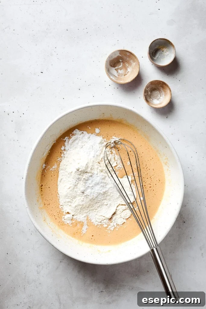 A mixture of dry ingredients including flour, cornstarch, baking soda, and salt is carefully added to the top of the wet blonde brownie batter in a mixing bowl.