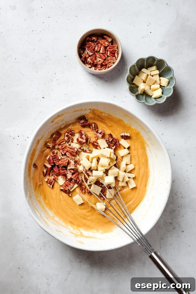 White chocolate chips and freshly chopped pecans are being stirred into a bowl of blonde brownies batter, ensuring even distribution of these delightful add-ins.