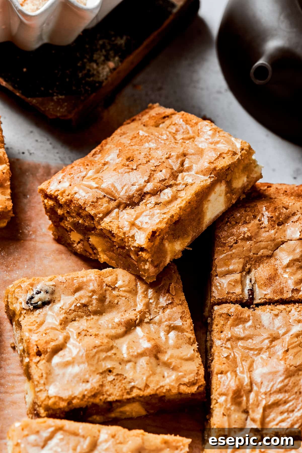 A single, perfectly baked blonde brownie square, showcasing its golden-brown crust, chewy texture, and visible white chocolate chips and pecans, rests atop a stack of more delicious brownies.