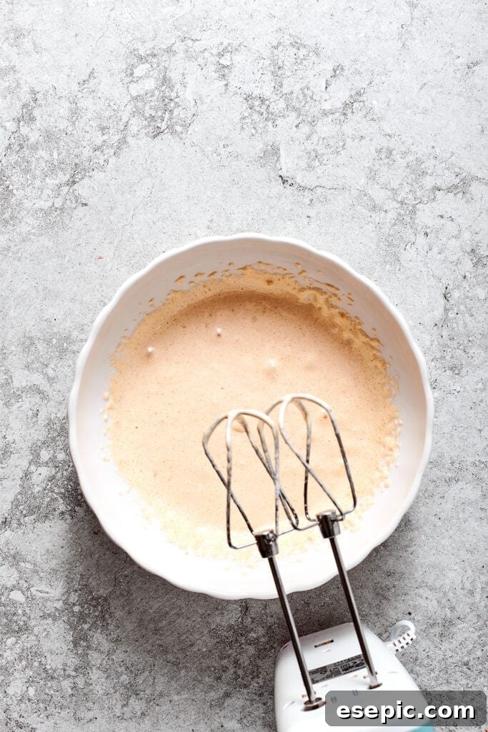 Beating the egg mixture until thick and cooled. A hand mixer is poised to beat the warmed egg mixture in a bowl, after it has been removed from the heat.