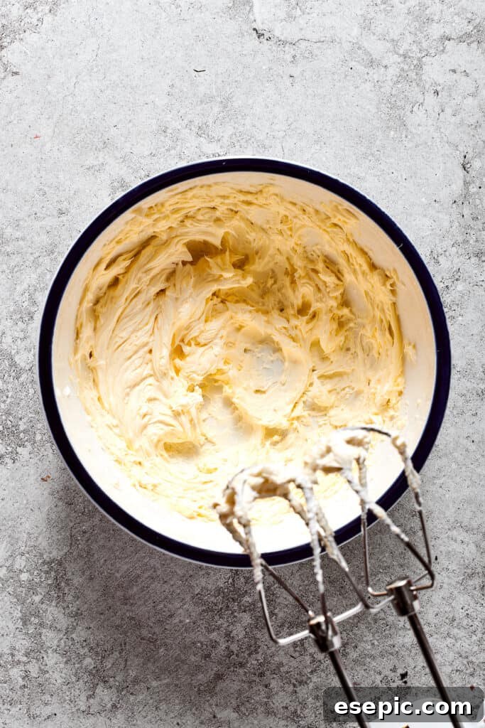 Whipping the butter until light and fluffy. Softened unsalted butter is being whipped to a light and fluffy consistency with a hand mixer in a separate bowl.
