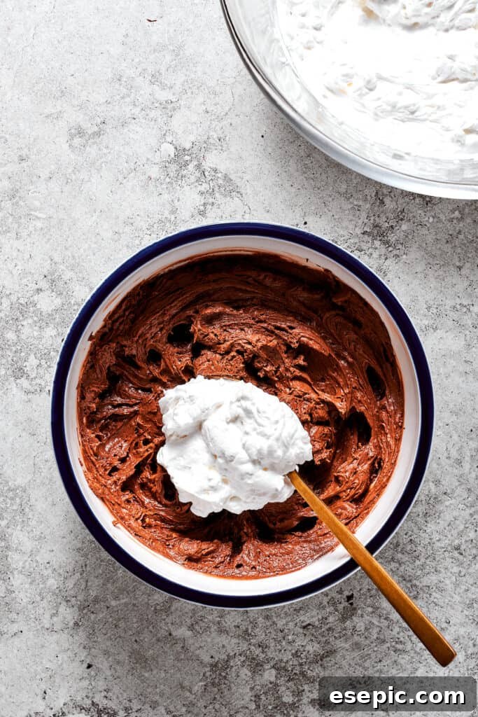 Gently folding whipped cream into the chocolate mixture. A spoonful of whipped cream is being gently folded into the chocolate filling in a bowl, demonstrating the delicate blending technique.