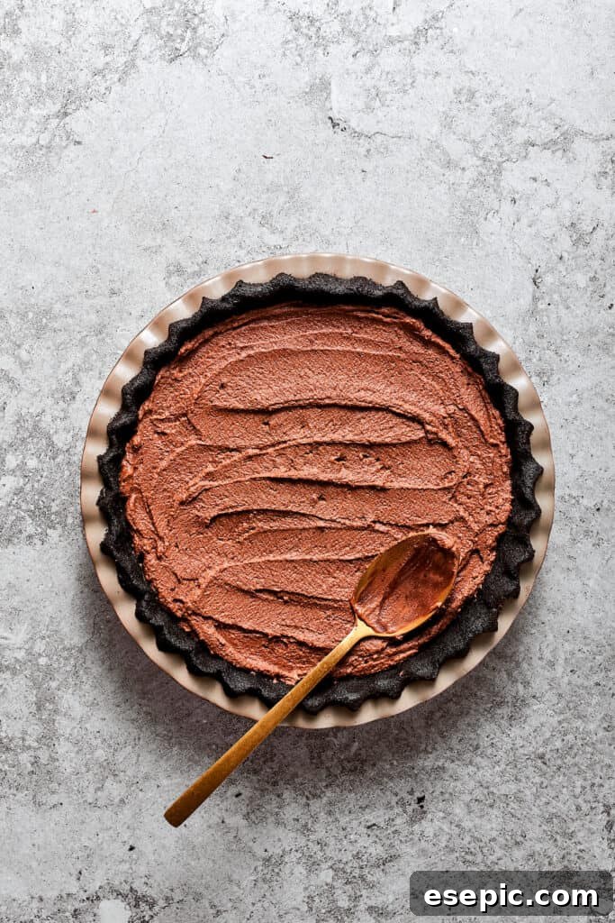 Filling the prepared pie crust with the chocolate mixture. A spoon smoothing the rich chocolate French Silk Pie filling into the chilled Oreo cookie crust in a pie dish.