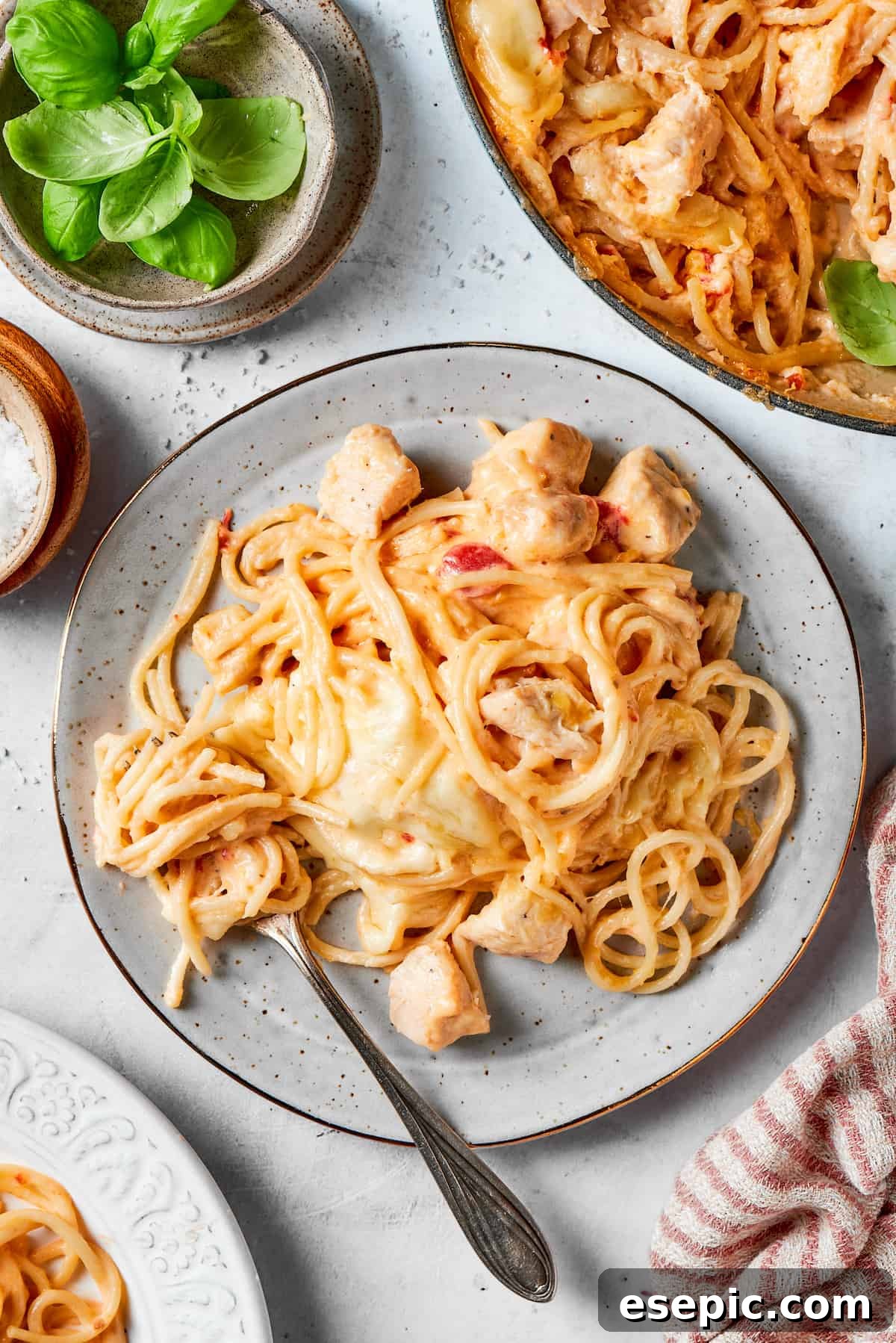 Hearty serving of Cheesy Chicken Spaghetti A plate of cheesy chicken spaghetti with a fork and a bowl of the spaghetti to the right, garnished with fresh basil.