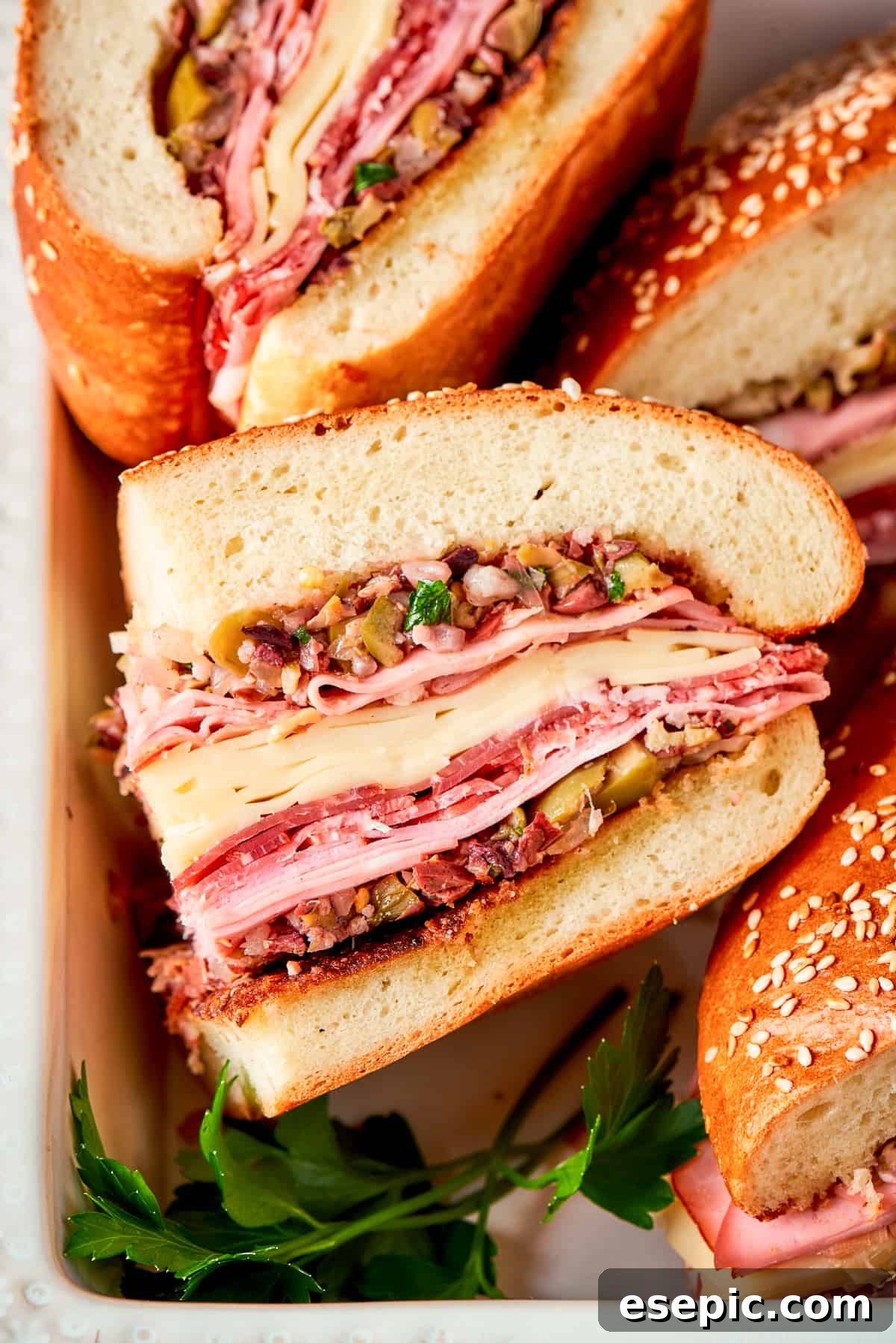 Two halves of a generous Muffaletta sandwich are displayed, showcasing the rich layers of meats, cheeses, and bright olive salad from a side angle.