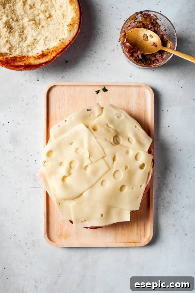 Slices of Swiss cheese are meticulously placed on top of the deli meats, forming a creamy layer in the muffaletta sandwich.