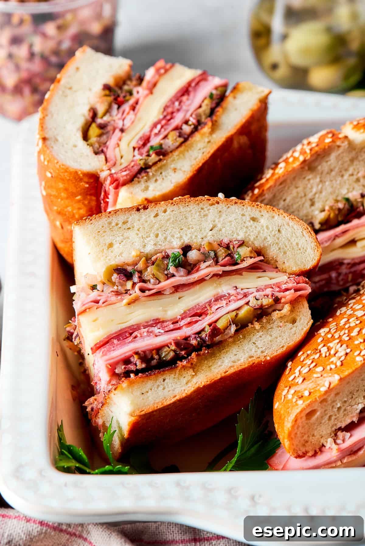Individual portions of a thick, layered Muffaletta sandwich are arranged on a wooden board, with all the delicious meats, cheeses, and olive salad clearly visible from the side.
