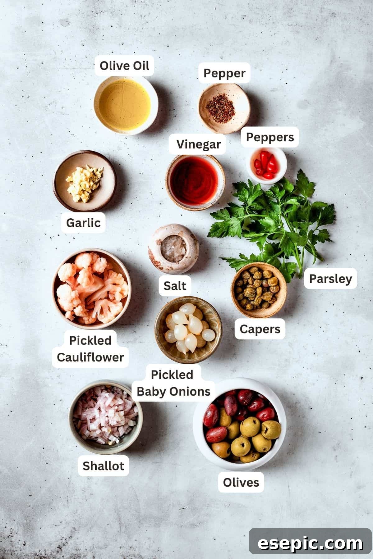 A vibrant selection of ingredients for the homemade olive salad, including green Castelvetrano olives, dark Kalamata olives, capers, Piri Piri peppers, shallots, garlic, fresh parsley, giardiniera, pickled baby onions, red wine vinegar, and olive oil.