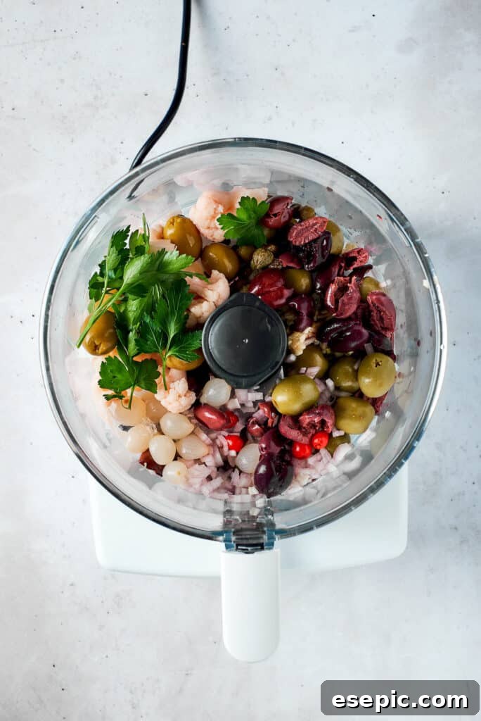 Various olive salad ingredients, including olives, peppers, and pickled vegetables, are being added to a food processor bowl.