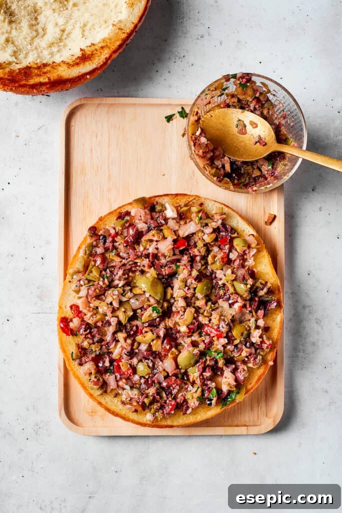 A generous layer of vibrant homemade olive salad is spread evenly onto one toasted half of the muffuletta bread.