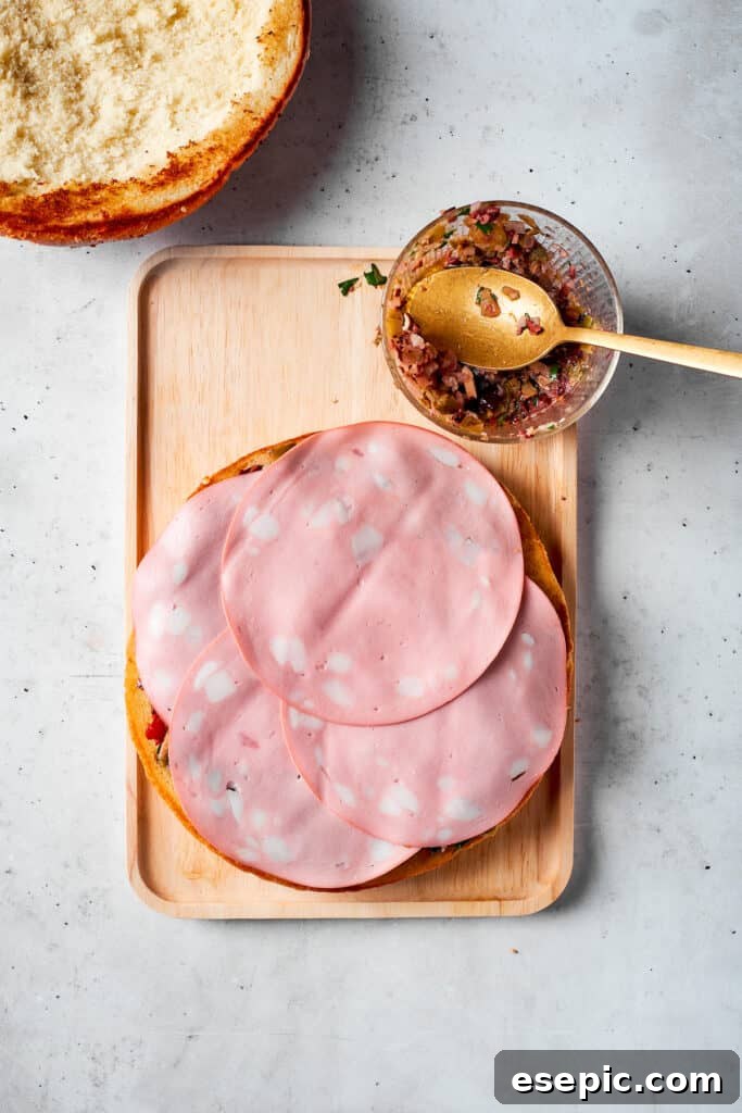 The final pieces of mortadella are delicately placed on top of the already stacked deli meats and cheeses, completing a layer of the muffaletta sandwich.