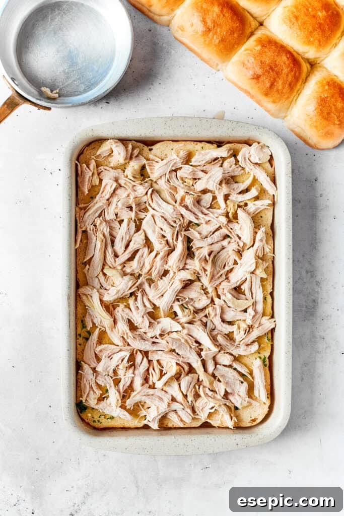 Shredded chicken being added to the bottom layer of Hawaiian rolls in a baking pan for the sliders.