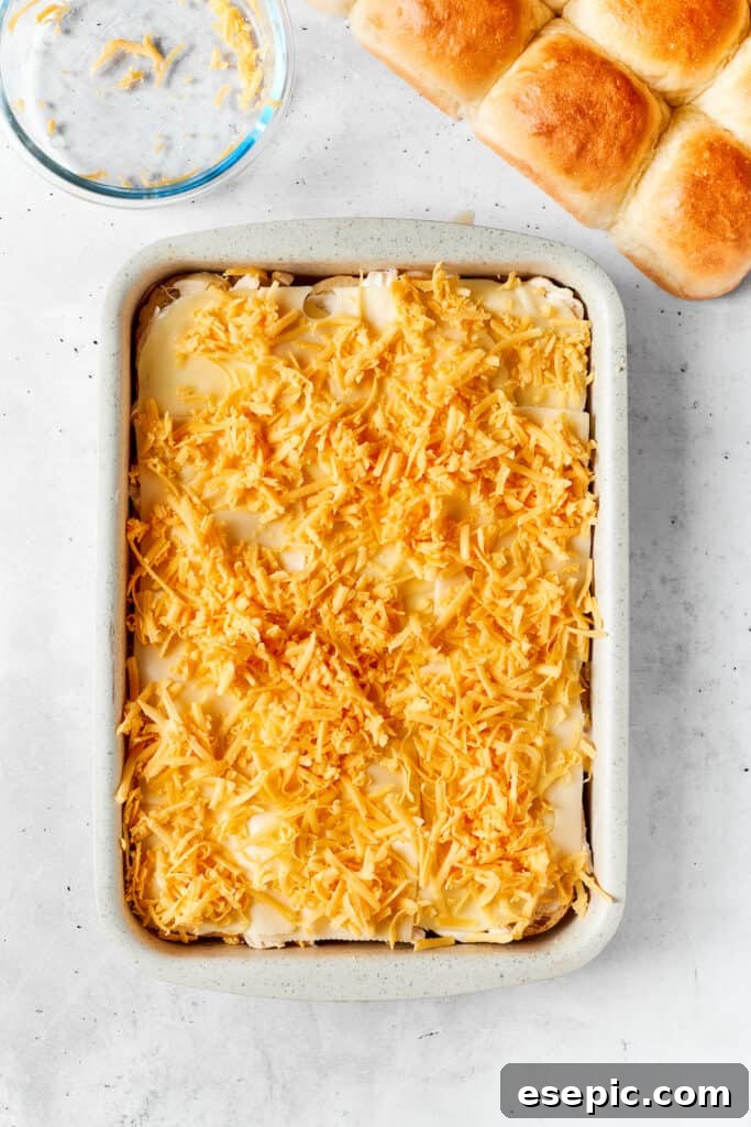 Shredded cheddar cheese being spread generously over the Provolone layer on the sliders.