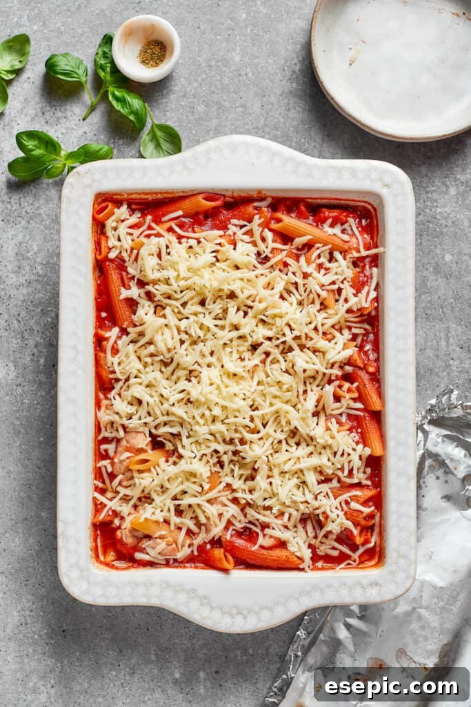 Shredded mozzarella cheese being generously sprinkled over the hot, partially baked chicken parmesan casserole.