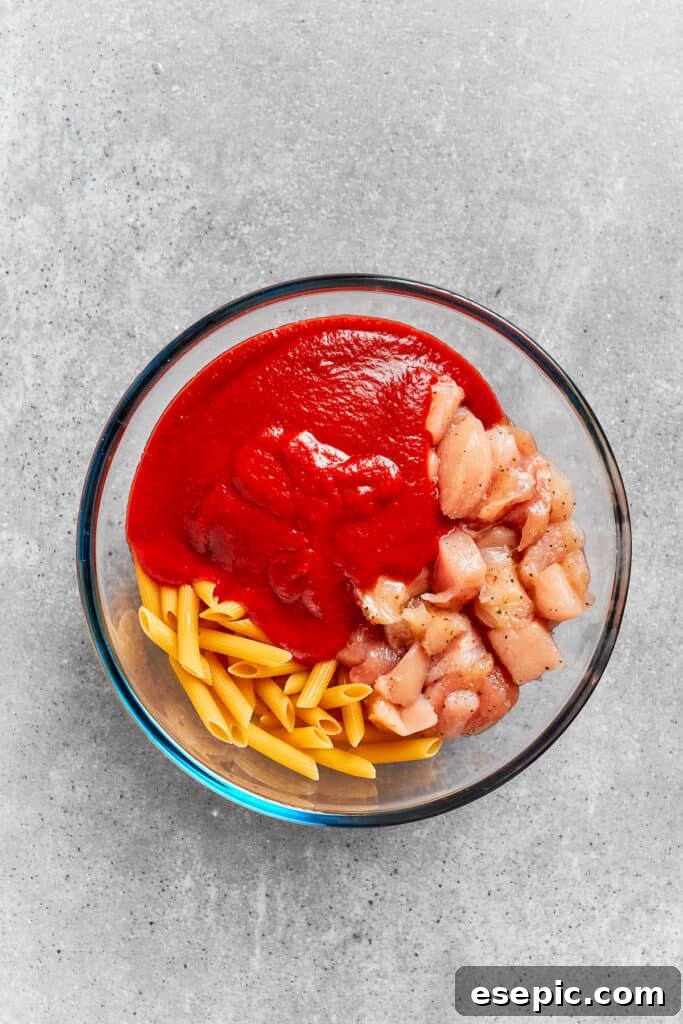 Rich marinara sauce being poured over the penne pasta and diced chicken in a mixing bowl.