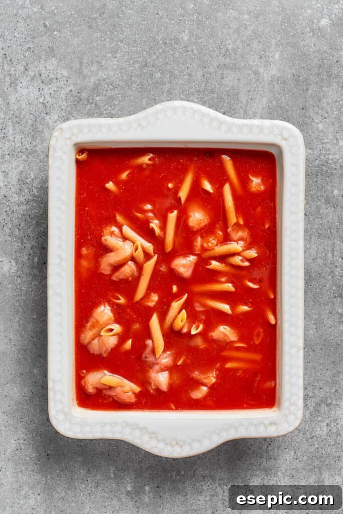 The mixture of uncooked penne, chicken, and marinara sauce being transferred from a bowl into a greased baking dish.