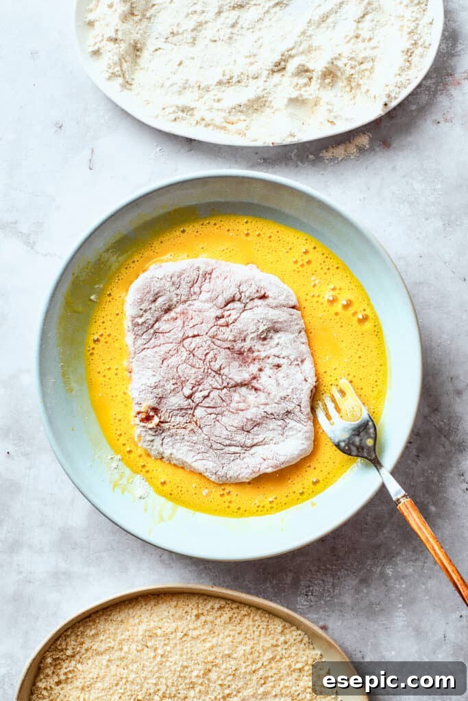 Dredging the pork chops in flour and eggs. A pork cutlet is carefully dipped into the beaten egg mixture, the second step in the breading process.