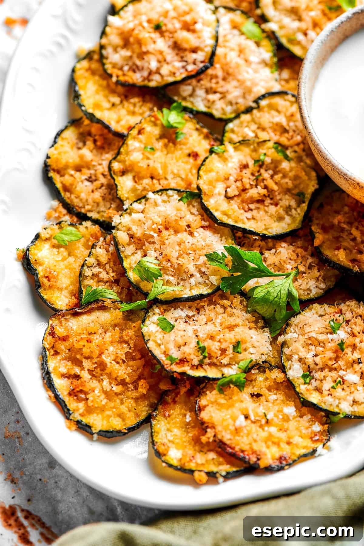 A pile of golden, crispy zucchini chips arranged on a pristine white plate, garnished lightly with fresh green herbs.