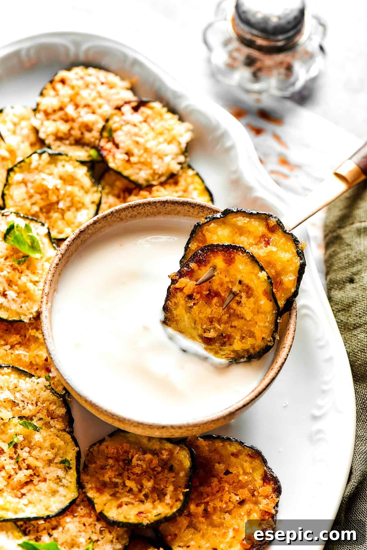 Two crispy zucchini chips delicately placed in a small bowl of creamy yogurt, surrounded by more chips on an elegant serving platter.