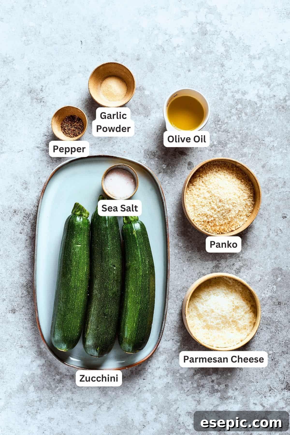 Ingredients for zucchini chips are neatly displayed and labeled: fresh zucchini, garlic powder, black pepper, olive oil, Panko breadcrumbs, sea salt, and Parmesan cheese.
