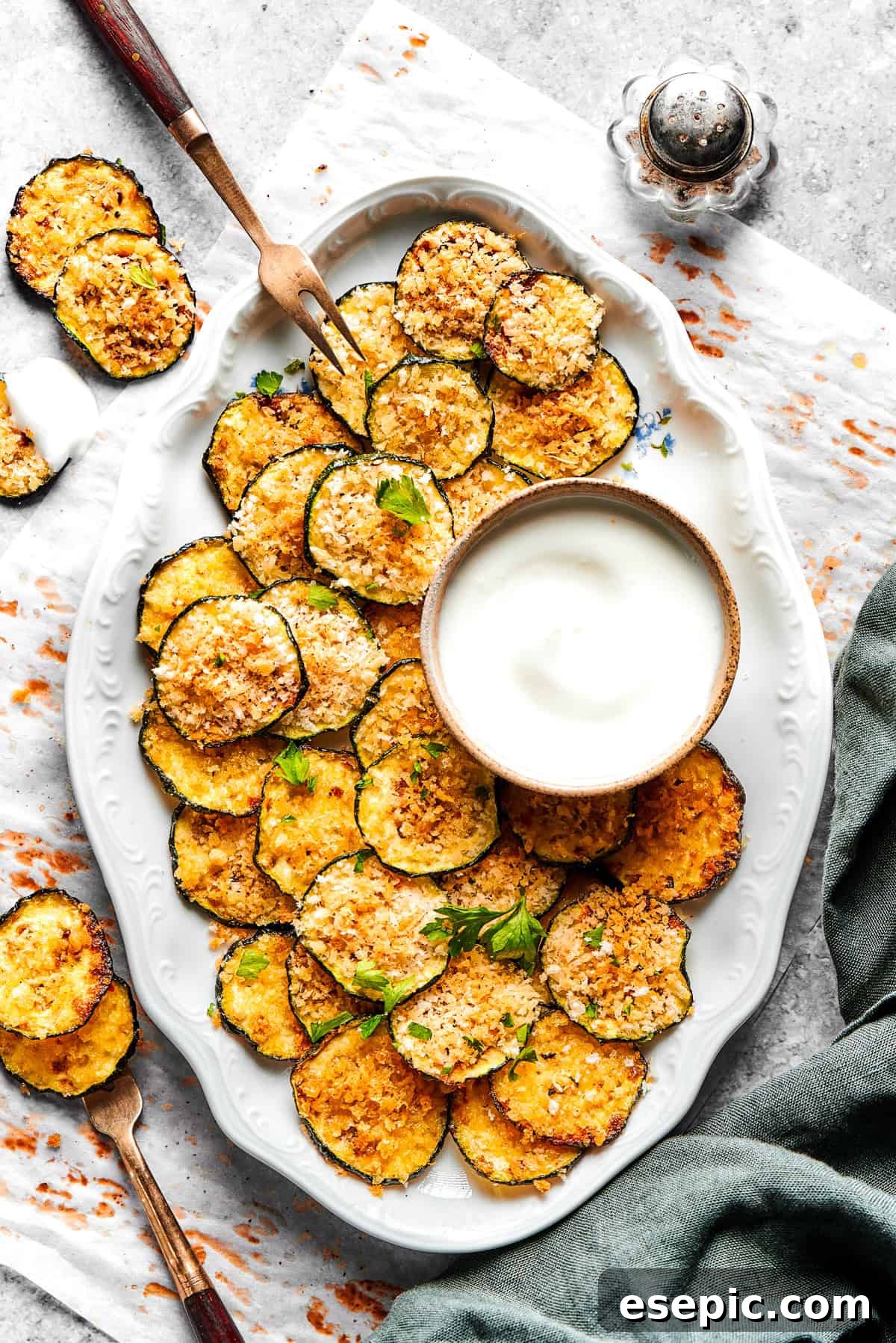 A serving of perfectly baked zucchini chips, arranged elegantly on an oval white platter with a small bowl of creamy dip on the side.