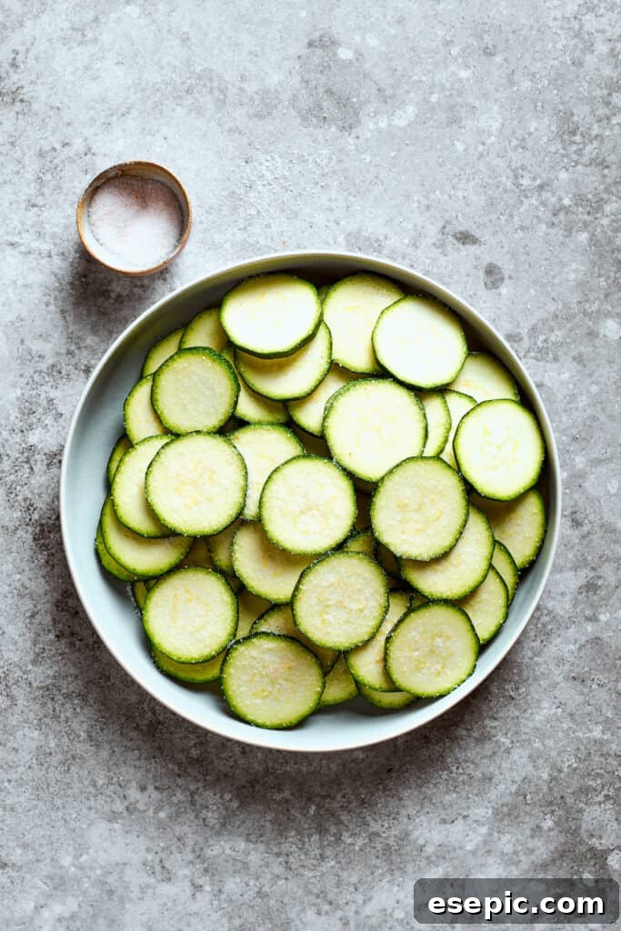 A bowl filled with uniformly sliced zucchini rounds sits next to a small bowl of salt, ready for the next step of preparation.