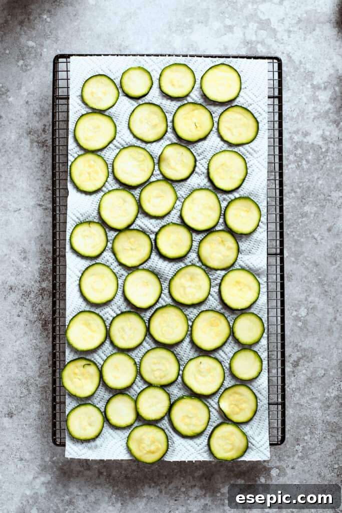 A large baking sheet is covered with a single layer of zucchini slices, lightly seasoned, before baking.