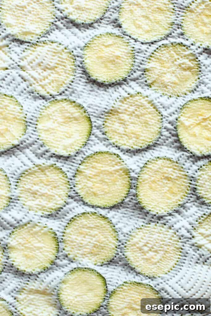 A baking sheet of zucchini slices is covered with a paper towel, illustrating the important step of patting them dry to remove excess moisture.