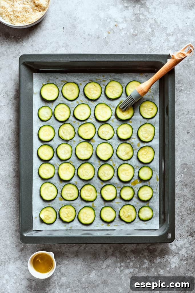 A pastry brush expertly applies olive oil to zucchini chips spread on a baking sheet, ensuring an even coating.