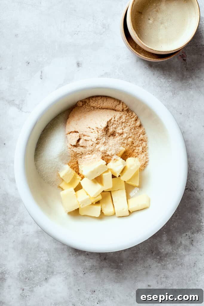 Next, for your chewy, buttery White Chocolate Chip Cookies, combine butter and sugars. Softened butter, granulated sugar, and brown sugar are added to a large white mixing bowl, ready to be creamed together for the cookie dough base.