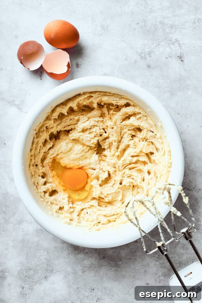 In the process of making chewy, buttery White Chocolate Chip Cookies, an egg is beaten into the butter-sugar mixture. A cracked egg is being added to the creamed butter and sugar mixture in a white bowl, ready to be beaten in to form the cookie dough.