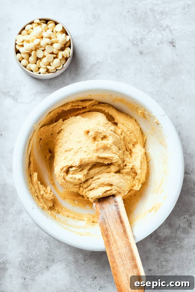 The cookie dough is stirred together for chewy, buttery White Chocolate Chip Cookies. The white chocolate chip cookie dough is being stirred in a large bowl, with all ingredients now combined to form a cohesive dough.