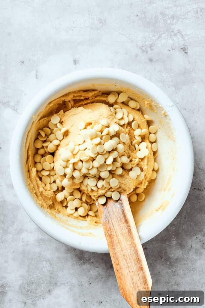White chocolate chips are added to the dough for chewy, buttery White Chocolate Chip Cookies. White chocolate chips are being gently folded into the prepared cookie dough in a large mixing bowl, ensuring even distribution.