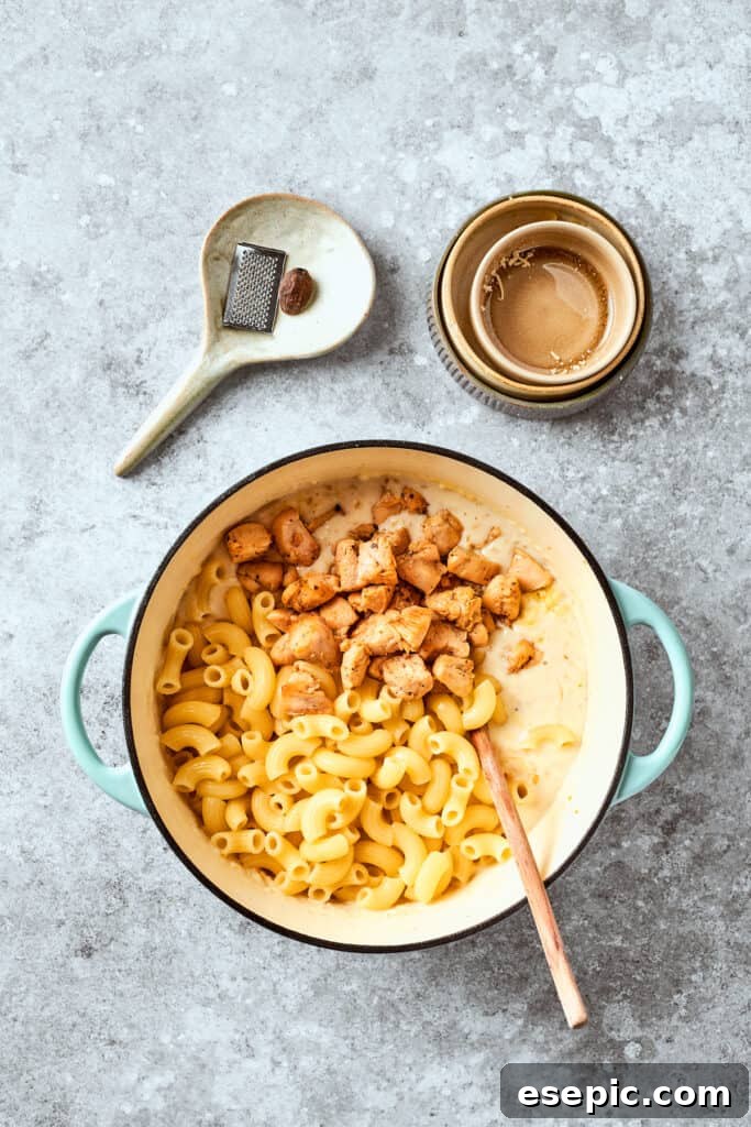 Cooked chicken being stirred into a pot of macaroni pasta and creamy cheese sauce.