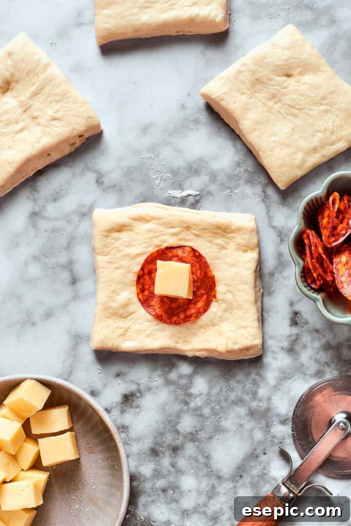 A slice of pepperoni and a cube of cheese is added to a square of dough.